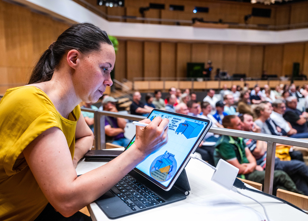 Eine Frau im gelben Shirt arbeitet an ihrem iPad, während einer Konferenz in einem großen Saal.