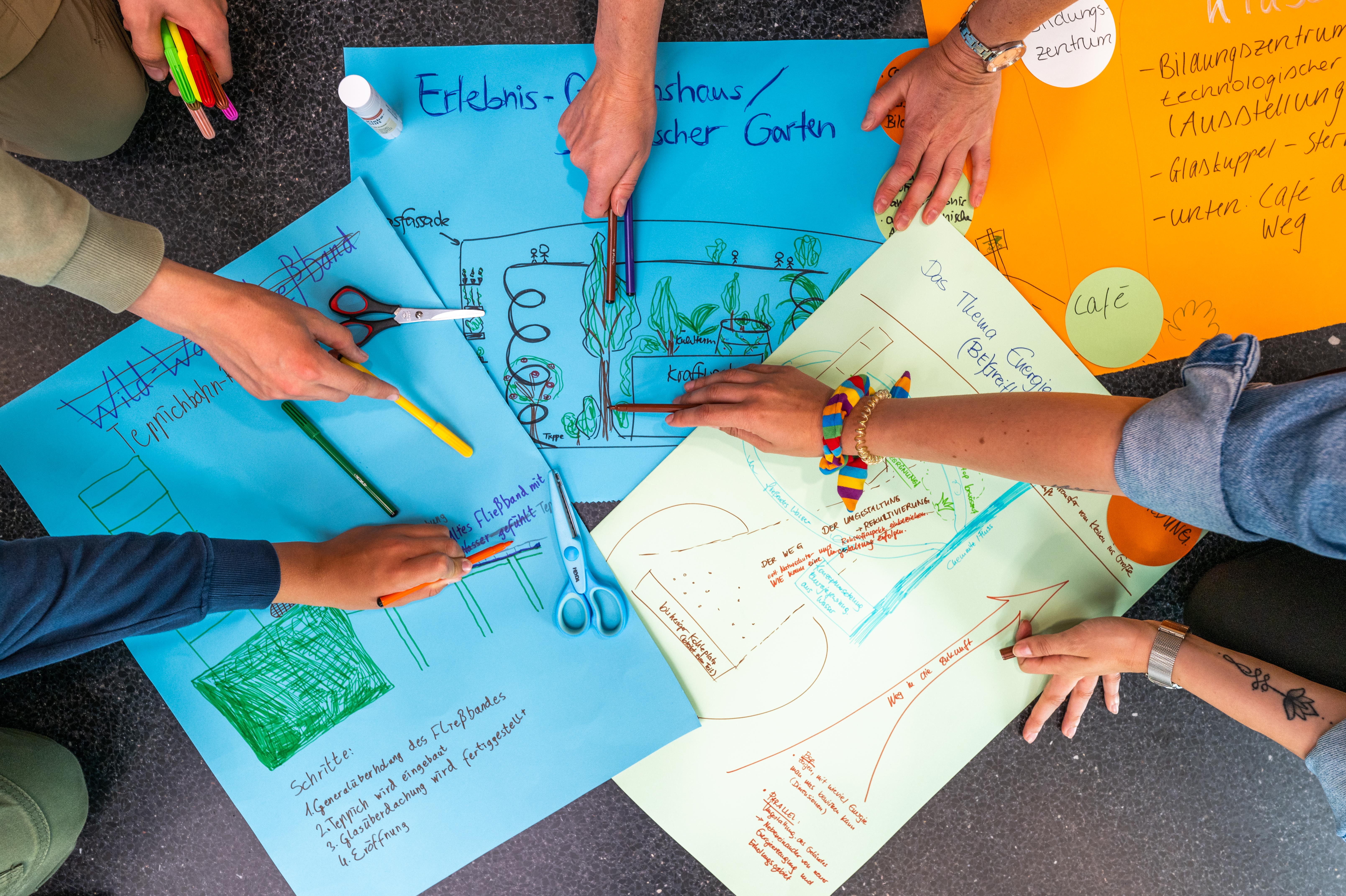 Mehrere Personen arbeiten gemeinsam an bunten Plakaten, die auf dem Boden liegen. Sie zeichnen und schreiben mit verschiedenen farbigen Stiften. Scheren und weitere Schreibutensilien sind ebenfalls zu sehen. Die Plakate zeigen Skizzen und Diagramme zu verschiedenen Energiethemen.