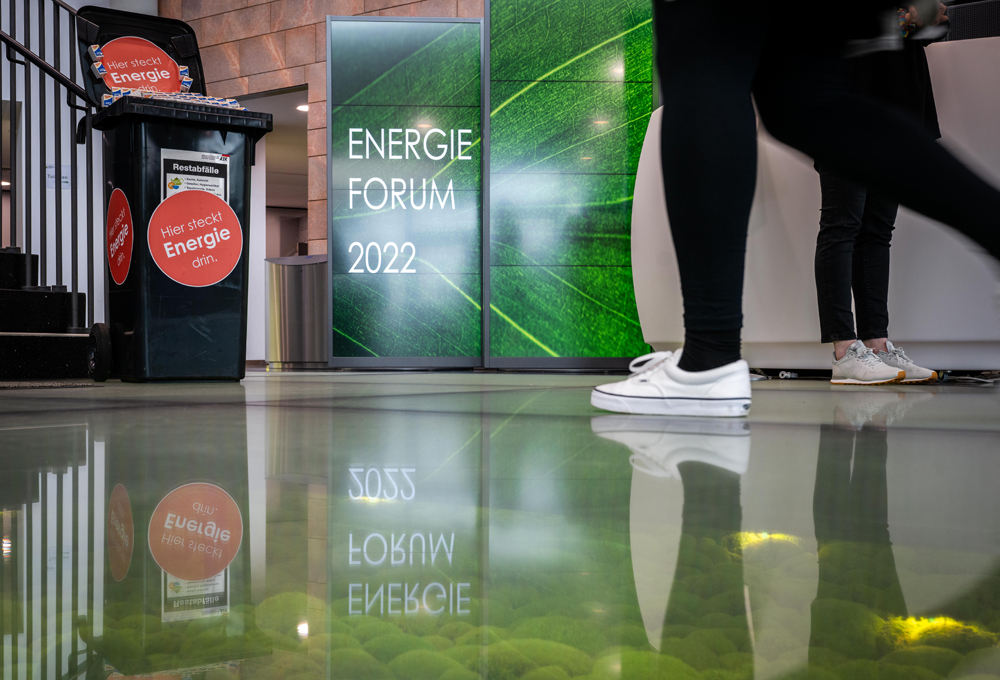 Eine Person läuft auf ein Schild mit der Beschriftung "Energieforum 2022" zu. Vor ihr steht ein Werbemittel in Form einer Mülltonne mit der Aufschrift "Hier steckt Energie drin".